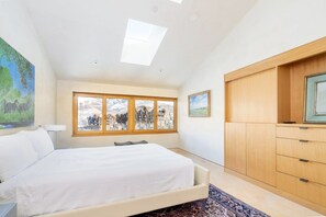 A bedroom with a bed, a window with a view of buildings, a skylight, and a wooden wardrobe.