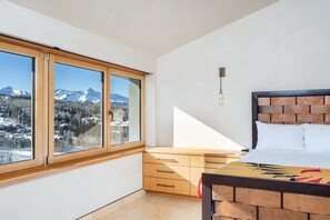 Un dormitorio con cama, cajones de madera, ventana con vista a la montaña y una lámpara.