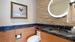 Een badkamer met een marmeren wastafel, houten kastjes, een toilet, een lijstwerk schilderij en een ronde spiegel met verlichting.