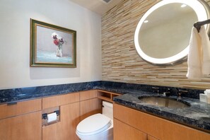Un baño con lavamanos de mármol, armarios de madera, inodoro, un cuadro enmarco y un espejo redondo con luz.
