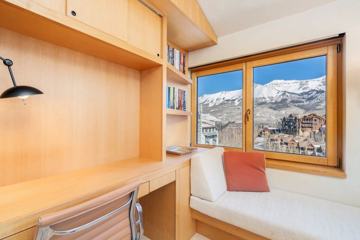 A room with a wooden desk, bookshelves, a cushioned bench, and a window with a snowy mountain view.