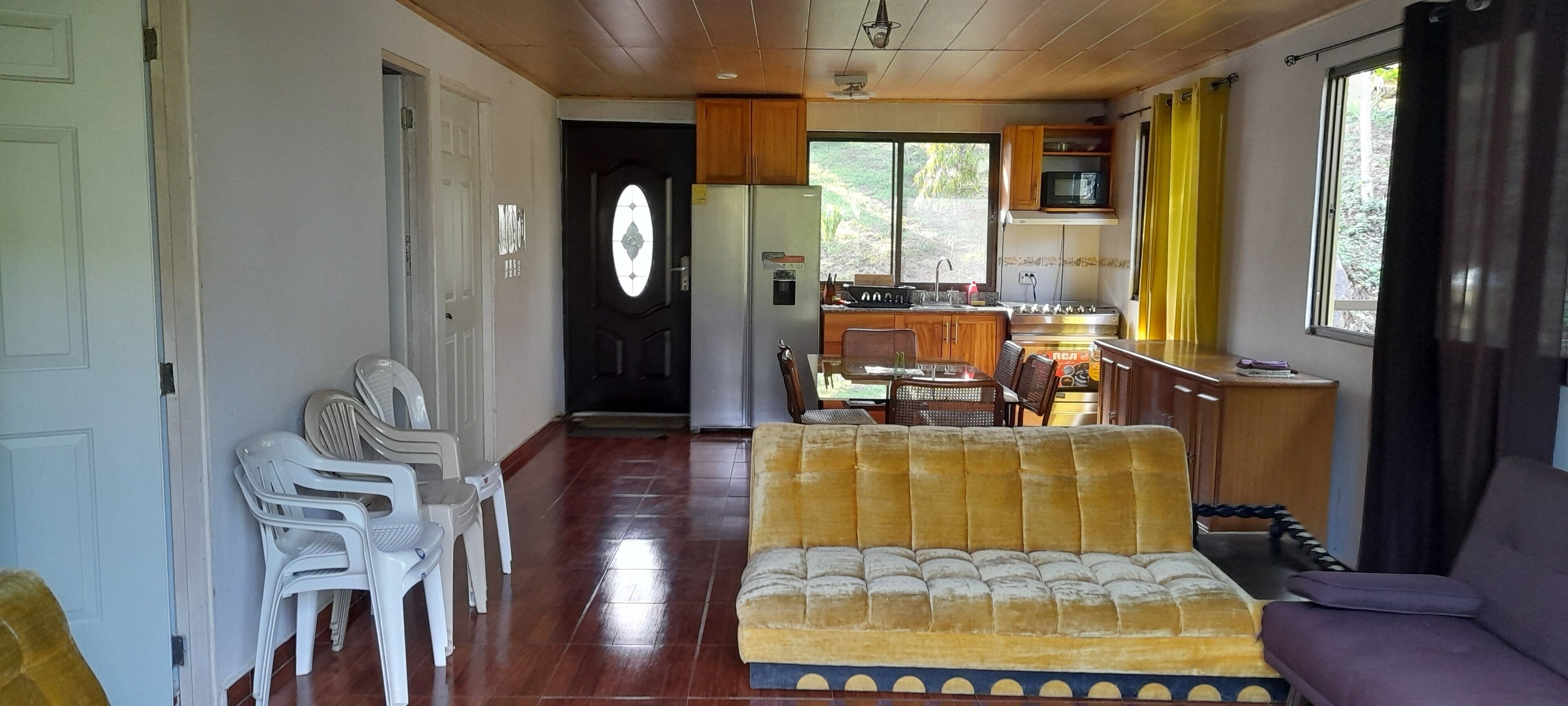 A living room with a yellow sofa, white plastic chairs, a dining table, and a kitchen area with wooden cabinets and a microwave.