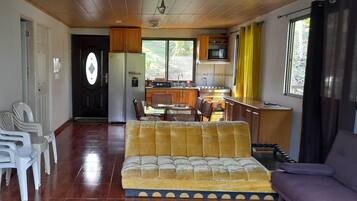 A living room with a yellow sofa, white plastic chairs, a dining table, and a kitchen area with wooden cabinets and a microwave.