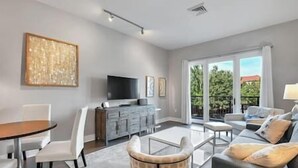 Ein modernes Wohnzimmer mit Essbereich, einer Sofagarnitur, einem Fernseher und einem großen Fenster mit Blick auf Grünflächen.