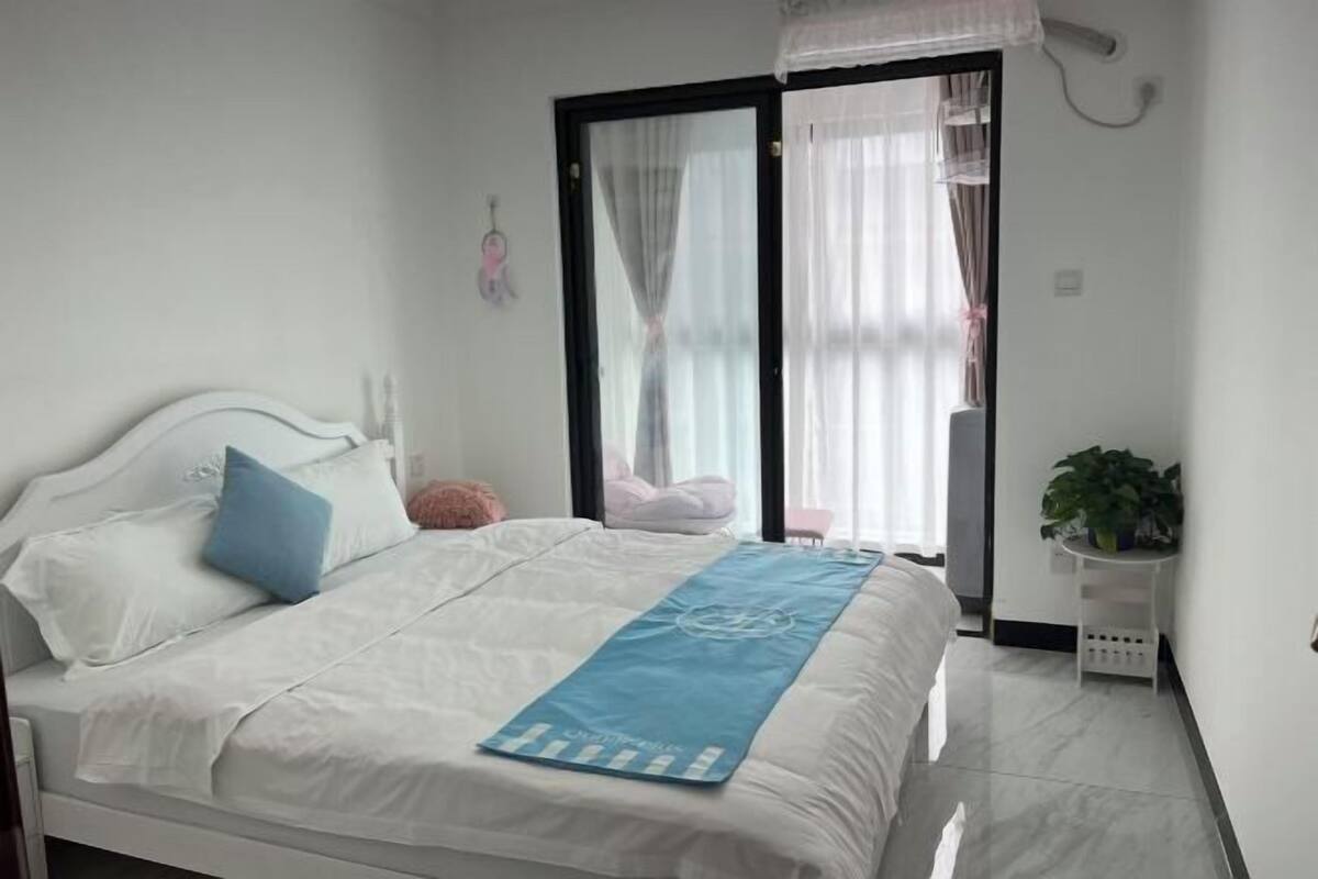 Quarto com cama, tapete azul, cabeceira branca, janela com cortinas, parede branca e pequena mesa branca com uma planta.