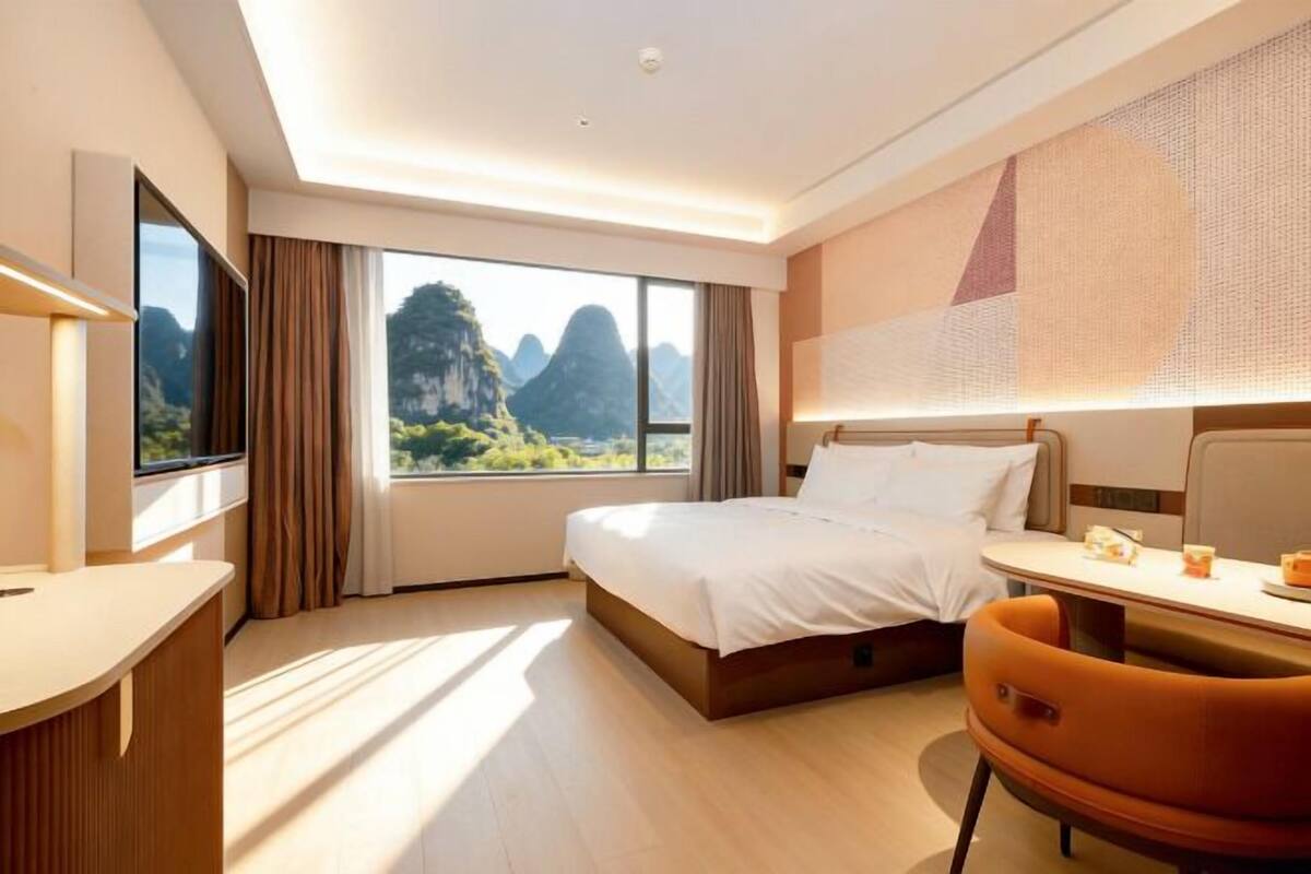 Quarto de hotel com cama, escrivaninha, cadeira, televisão e uma grande janela com vista para as montanhas.