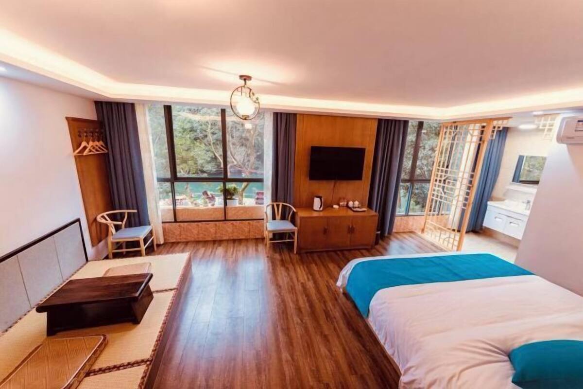 Quarto de hotel com cama, área de estar, televisão e janelas amplas.