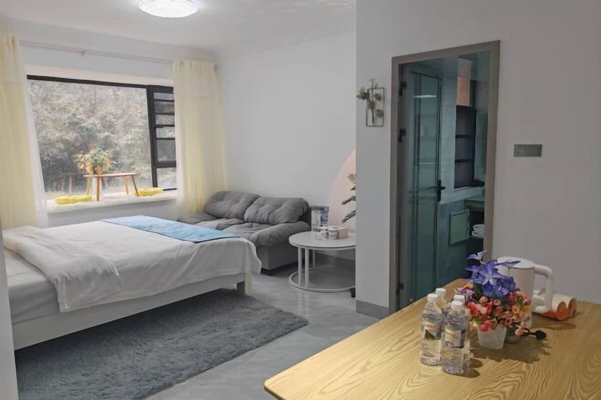 Um quarto moderno com uma cama, uma mesinha e uma escrivaninha. Há uma janela com cortinas, uma porta e uma vista de árvores lá fora.