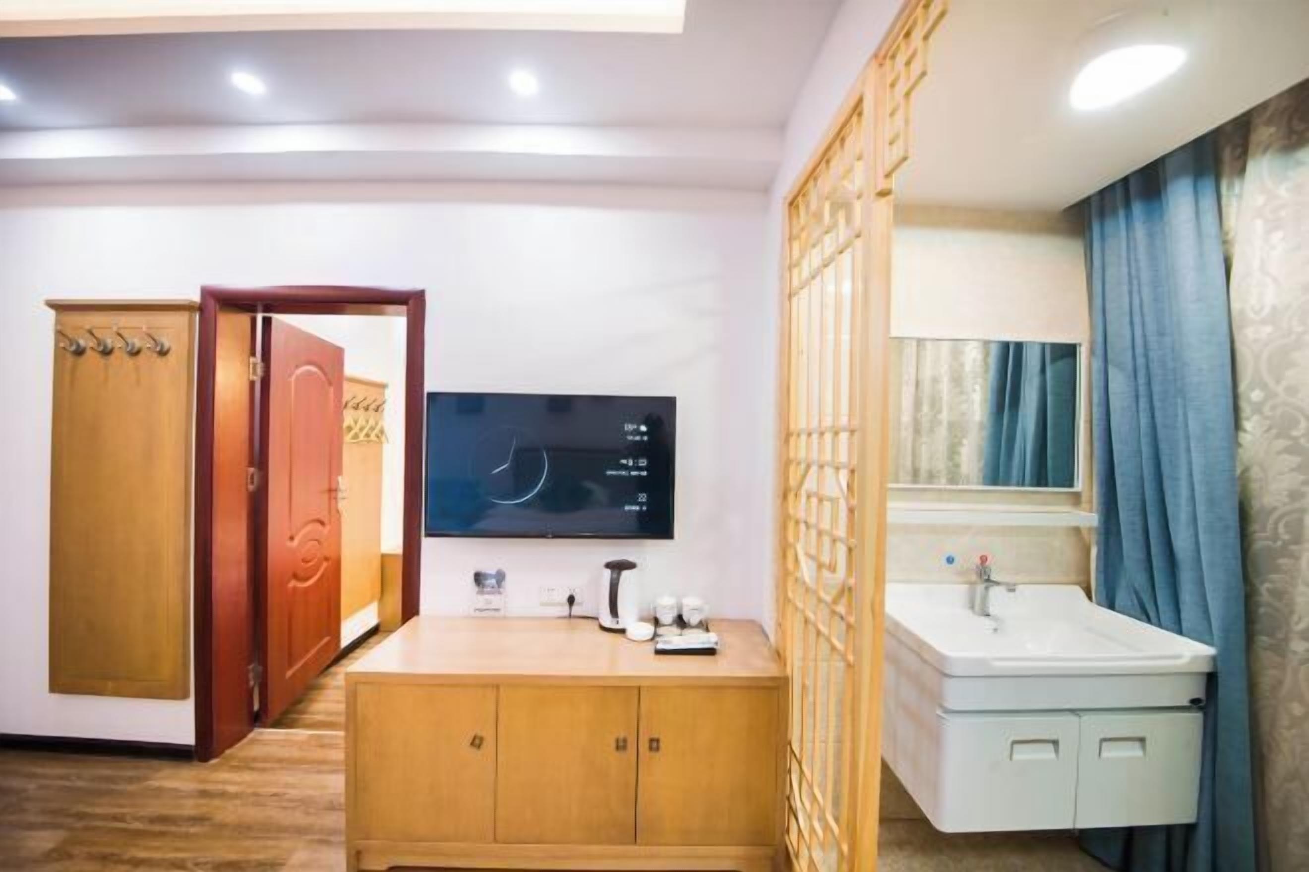 Une chambre d’hôtel avec une armoire en bois, un téléviseur fixé au mur, une table de chevet avec une lampe et une salle de bain équipée d’un lavabo et d’un miroir.