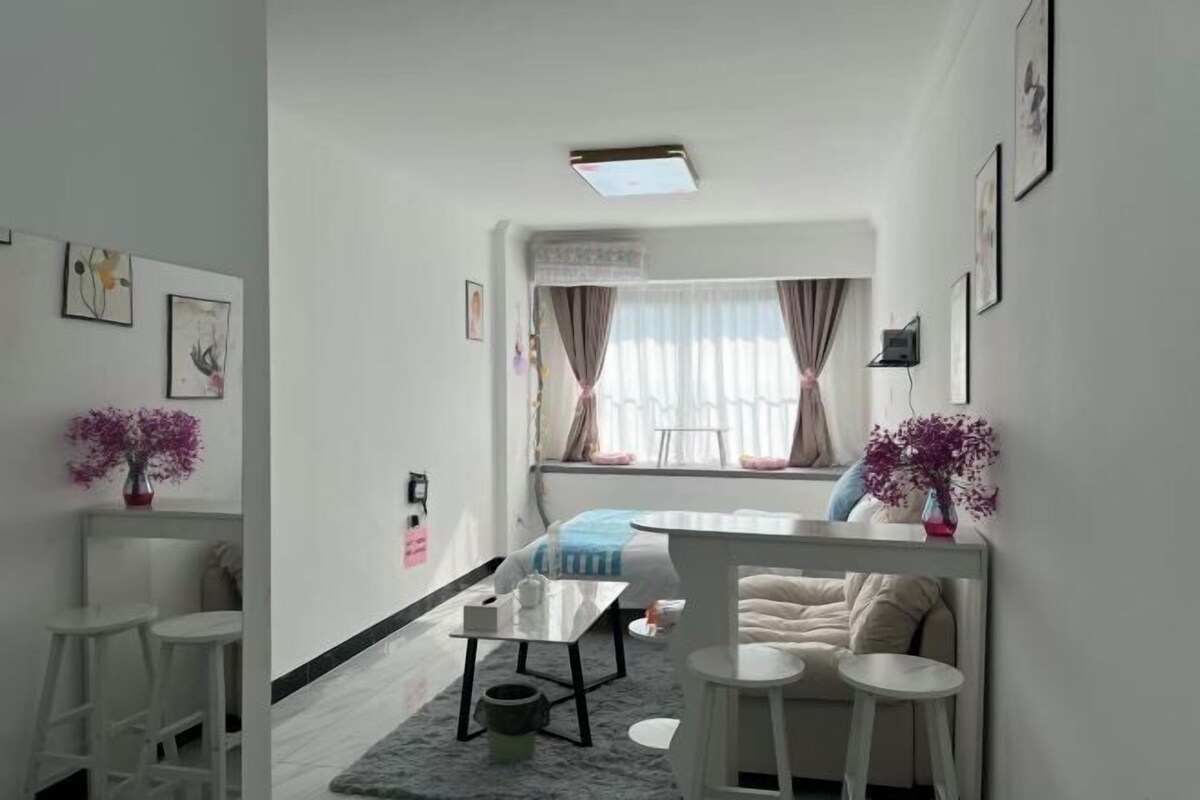 A modern, well-lit room with a bed, a small table, and two bar stools.
