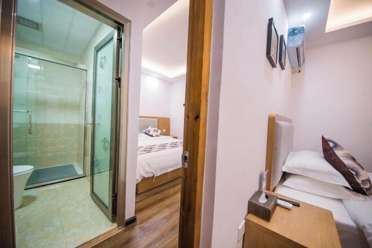 A hotel room with a glass-enclosed shower, a bed with a white comforter, a wooden bedside table, and a wall-mounted air conditioning unit.