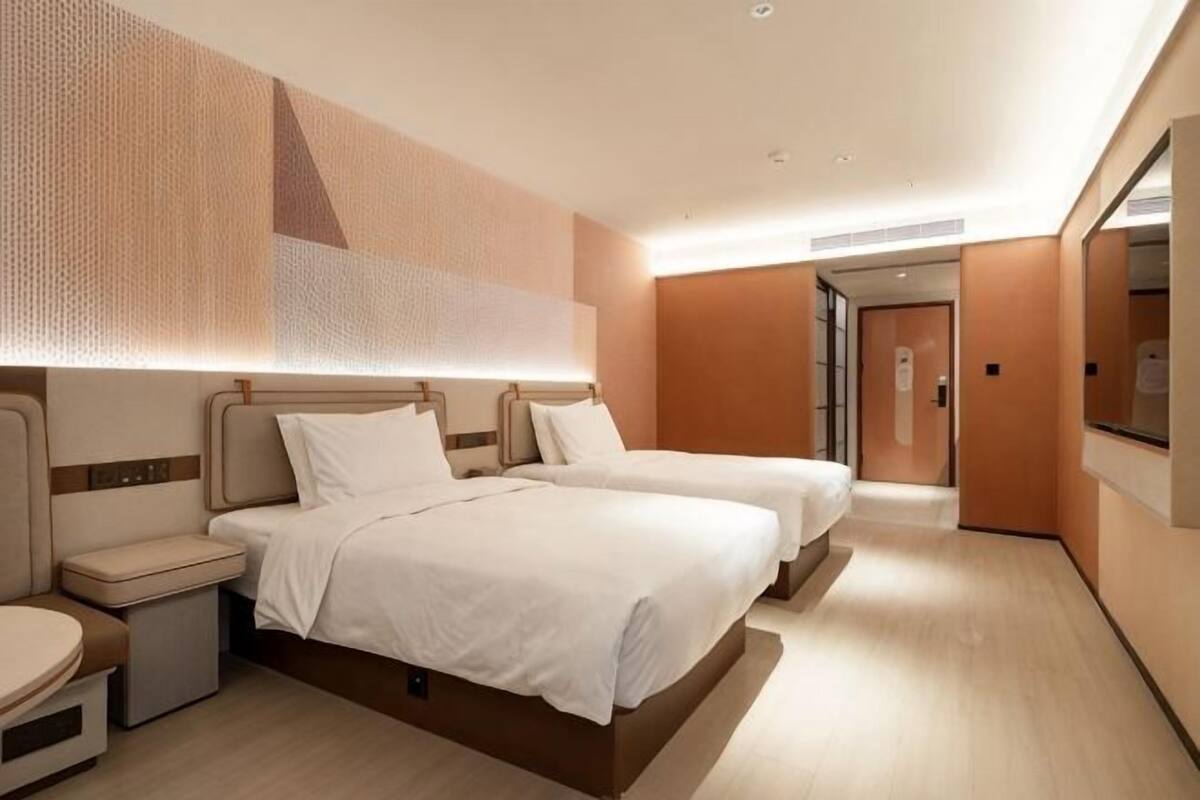 Quarto de hotel com duas camas, uma mesa de cabeceira, um espelho grande e uma porta que dá para outro quarto.