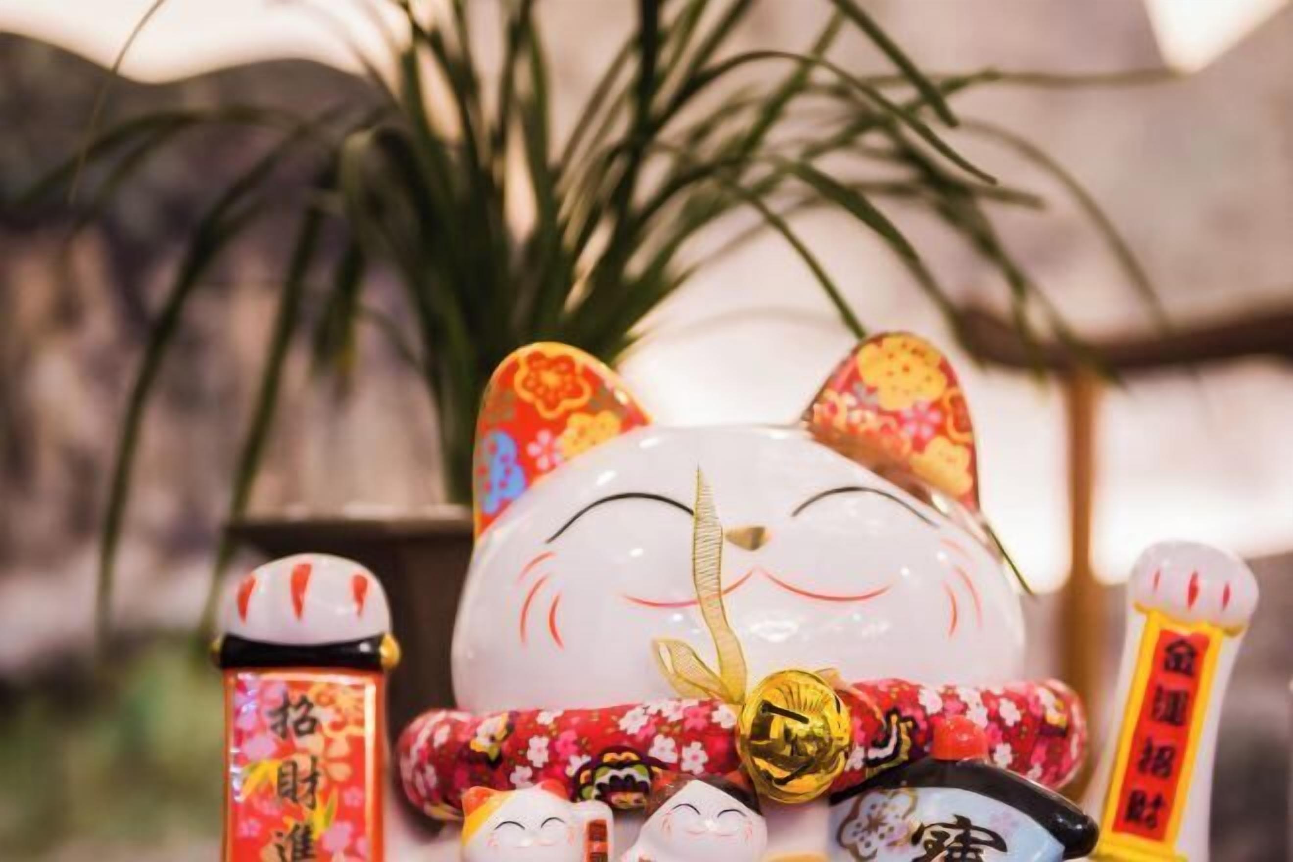 Eine dekorative Katzenfigur mit einer goldfarbenen Glocke und einem roten Band, umgeben von kleinen Figuren und einer Flasche mit japanischen Schriftzeichen.