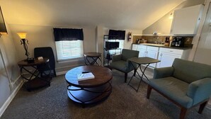 A compact living space with a round coffee table, two armchairs, a small side table, a lamp, a television, and a kitchenette.