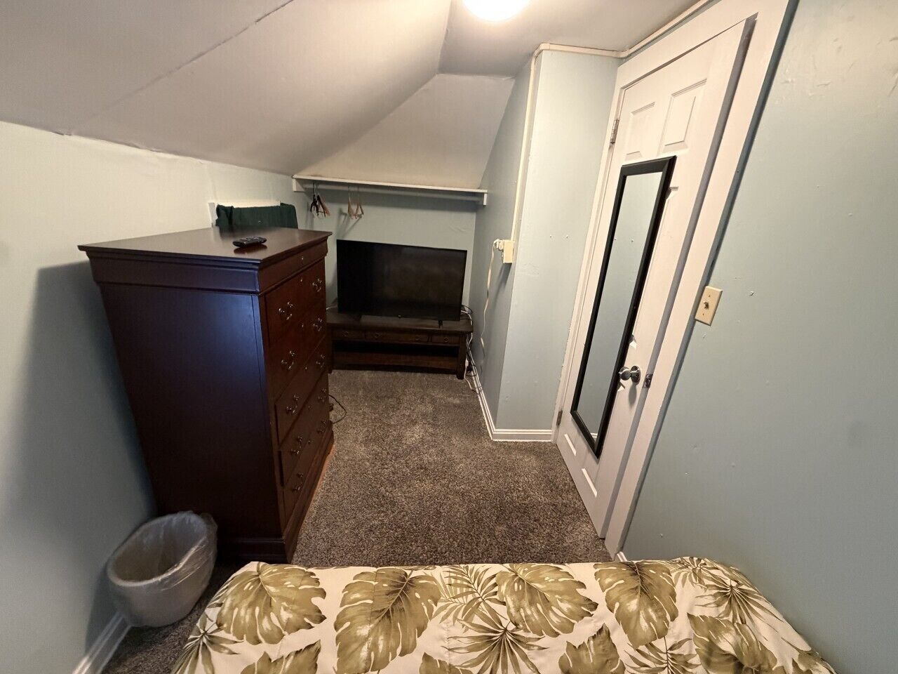 Une chambre à coucher avec une commode, un lit recouvert d’une couvre-lit à motifs floraux, une porte et une petite fenêtre.