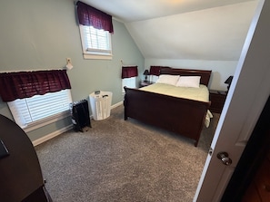 A bedroom with a bed, two nightstands, a window with curtains, a radiator, and a door.