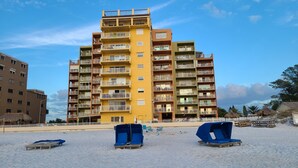 A multi-story building with balconies, a sandy beach in the foreground, and a clear sky. - Beachfront condo with intracoastal views in Tampa Bay area, heated-cooled pool! (Indian Rocks Beach)