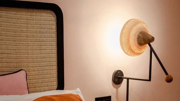 Ein Bett mit weißem und orangefarbenem Kissen, einem gewobenen Kopfteil und einer Wandlampe mit Holzfuß und runder Schirm.