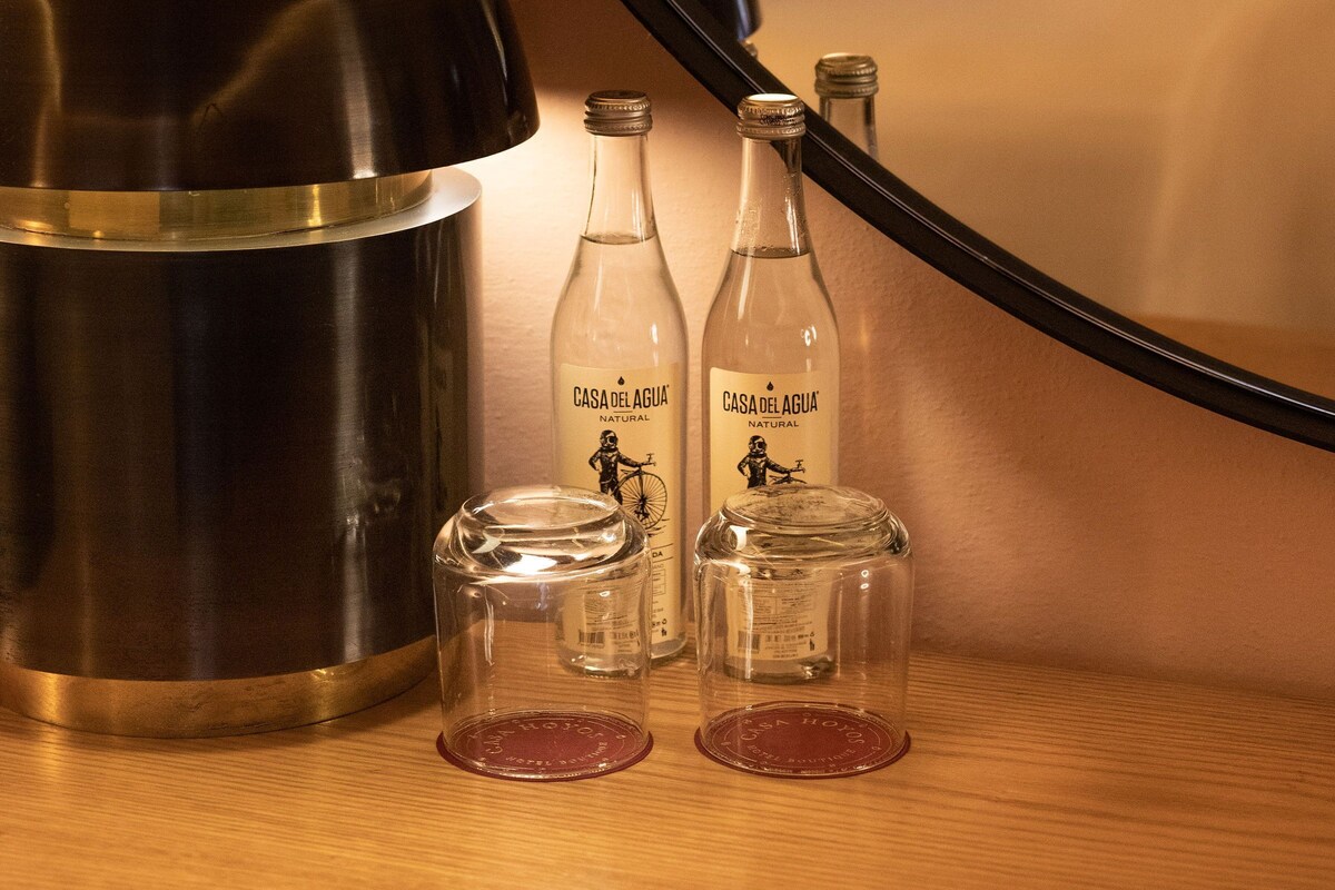 Ein Hotelzimmer mit einem Bett, einem Nachttisch mit Wasserkocher, zwei Wasserflaschen und einem Spiegel, der das Zimmer reflektiert.