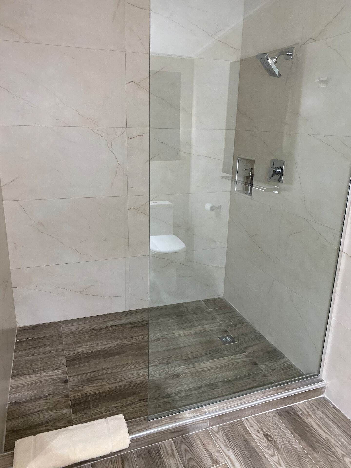 Una ducha con cerramiento de vidrio, piso de madera oscura y un inodoro blanco al fondo.
