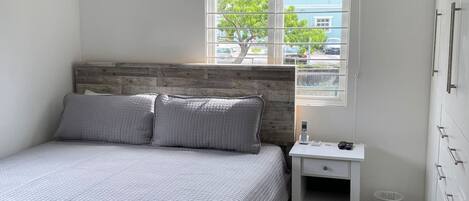 Un dormitorio con cama, mesita de noche, ventana con vistas a edificios y árboles, y un equipo de aire acondicionado.