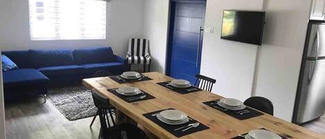 Ein modernes Wohnzimmer mit einem blauen Sofa, einem Esstisch mit Tellern und Besteck, einem gestreiften Sessel und einem an der Wand befestigten Fernseher.