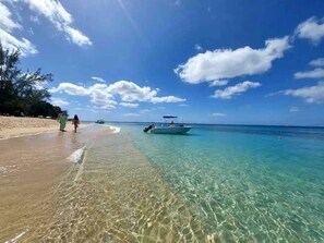 A beach with clear turquoise water, a boat, and a few people walking on the shore.