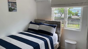 Un dormitorio con cama, aire acondicionado, ventana con vistas y un cubo de basura.