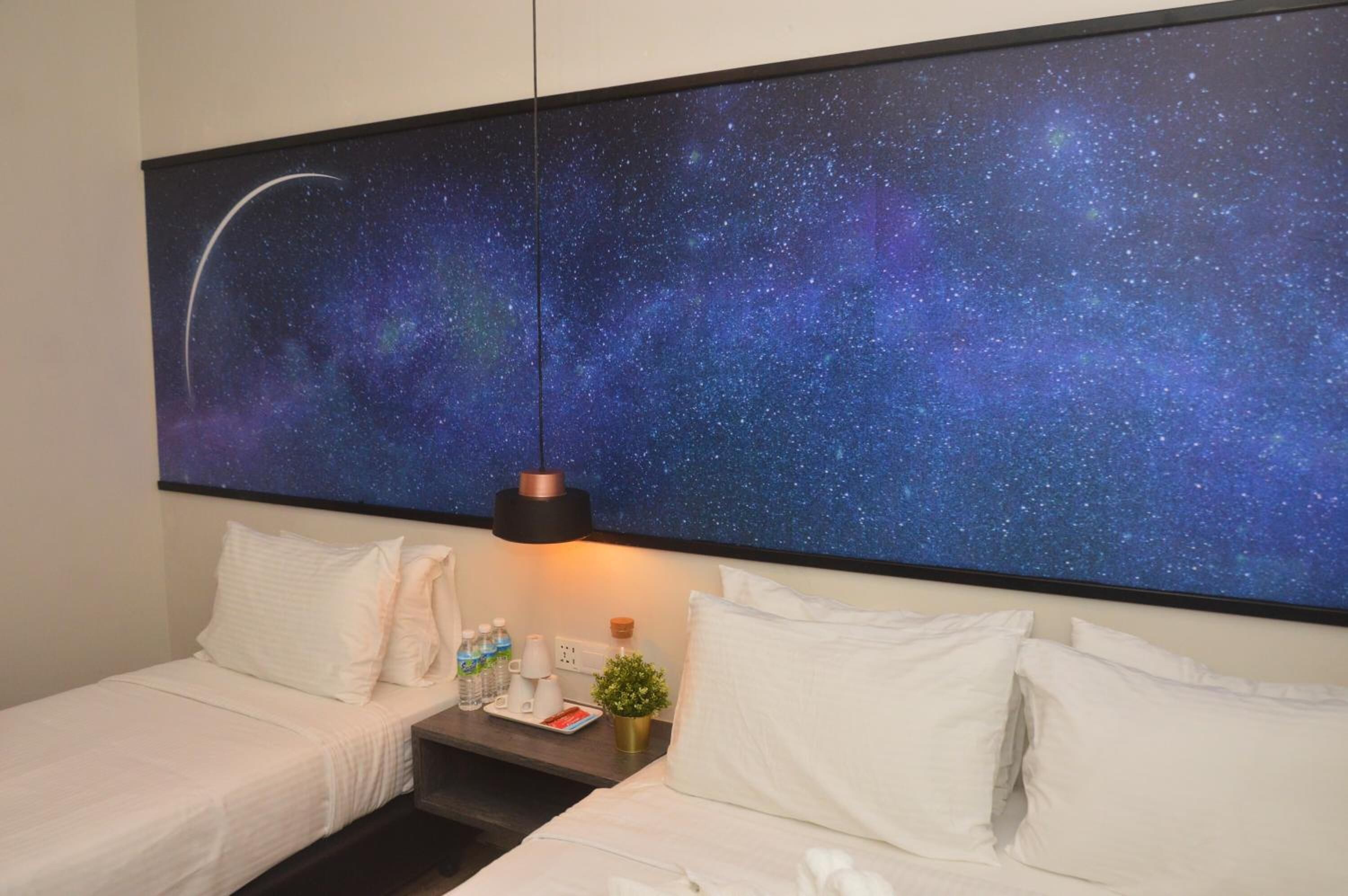Habitación de hotel con cama, mesita de noche, lámpara y un mural del cielo nocturno en la pared.