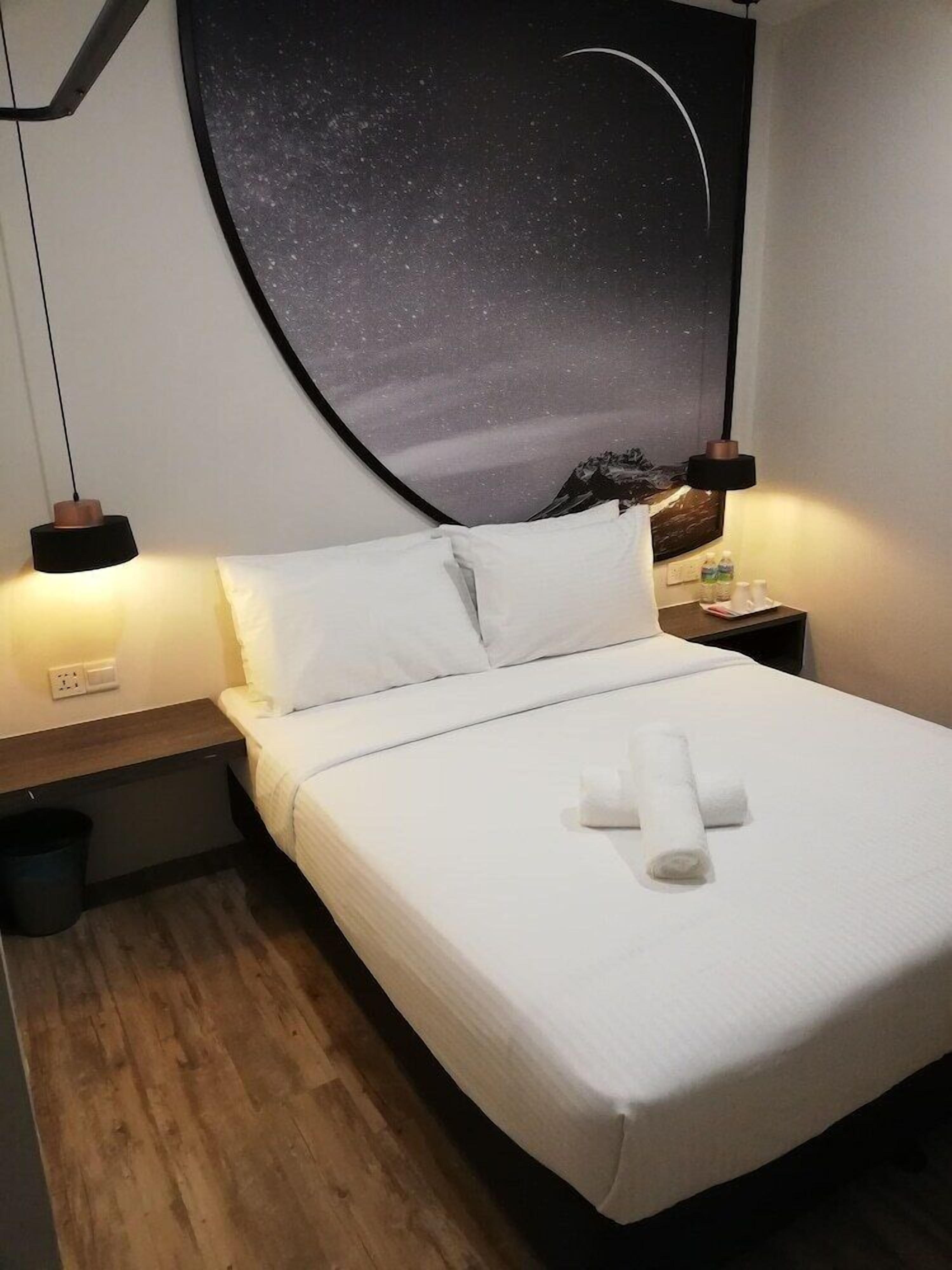 Una cama bien hecha con sábanas blancas y una toalla enrollada, una mesita de noche con una lámpara, y una pared con un mural del cielo estrellado.