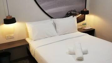 Una cama bien hecha con sábanas blancas y una toalla enrollada, una mesita de noche con una lámpara, y una pared con un mural del cielo estrellado.