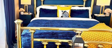Een slaapkamer met een blauwe wand, een gouden bed, een schilderij en een blauw kamerkleed.