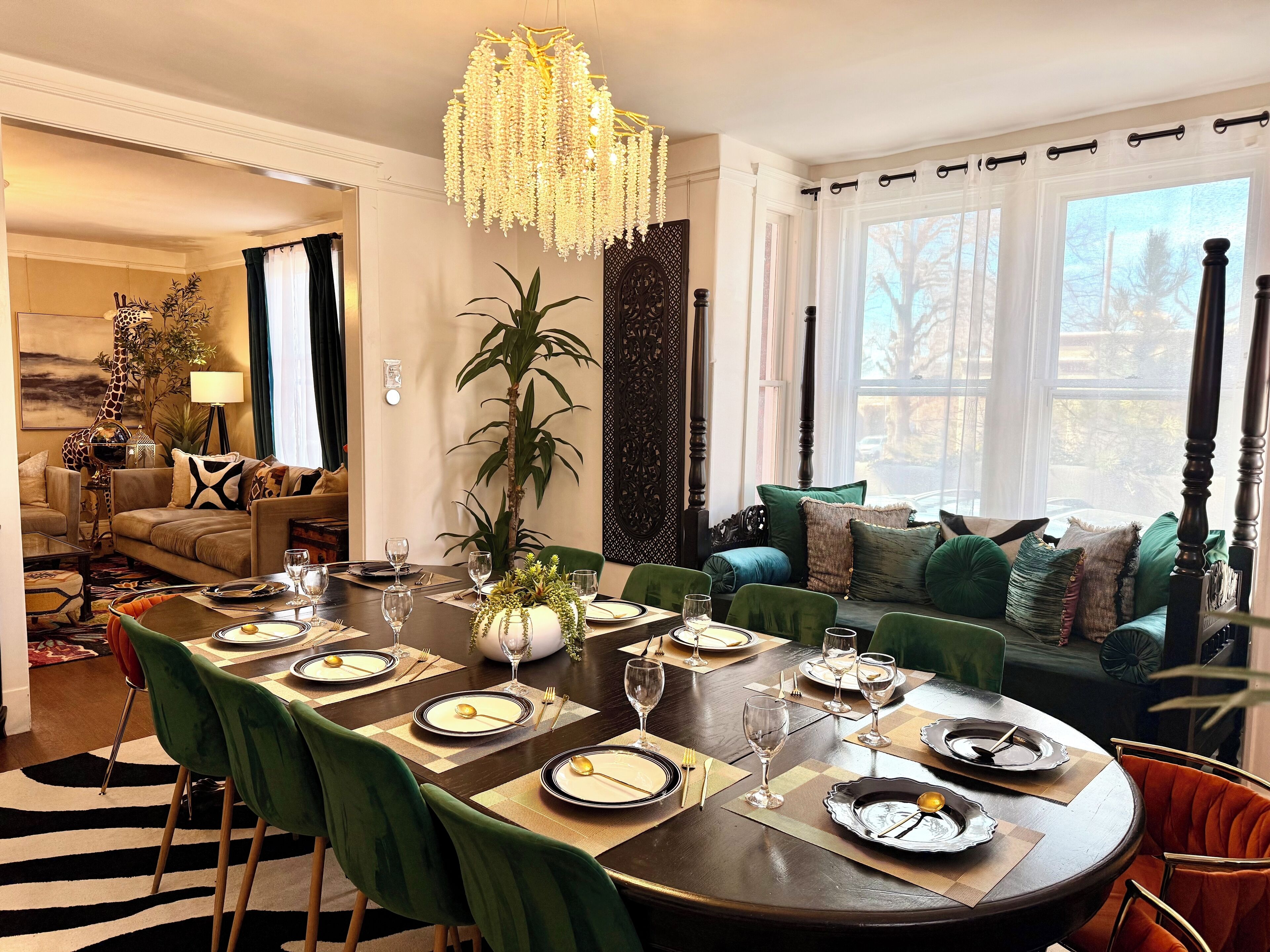 A dining room with a round table set for six, green upholstered chairs, and a large window with a view of trees.