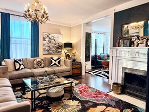 A living room with a sofa, coffee table, and fireplace. There is a chandelier, artwork on the wall, and a view into another room. - Elliot Barker Estate for Large Groups Stroll to Canyon Rd & Plaza (Santa Fe)