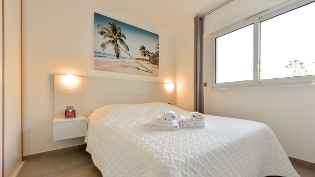 Una cama bien hecha con un edredón blanco y dos toallas enrolladas, un estante en la pared con un objeto pequeño, y un cuadro con motivos playeros sobre la cama.