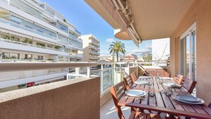 Un balcón con mesas y sillas de madera, una palmera y vista a un edificio moderno con balcones.