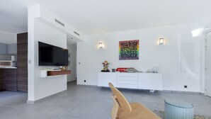 Una sala de estar moderna con una mesa de comedor de madera, un televisor montado en la pared, un armario blanco y un cuadro abstracto y colorido.