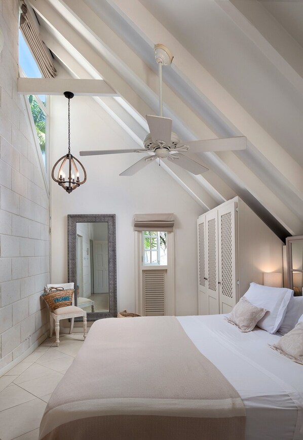A bedroom with a bed, a ceiling fan, a chandelier, a mirror, and a window with shutters, a door, and a wardrobe.