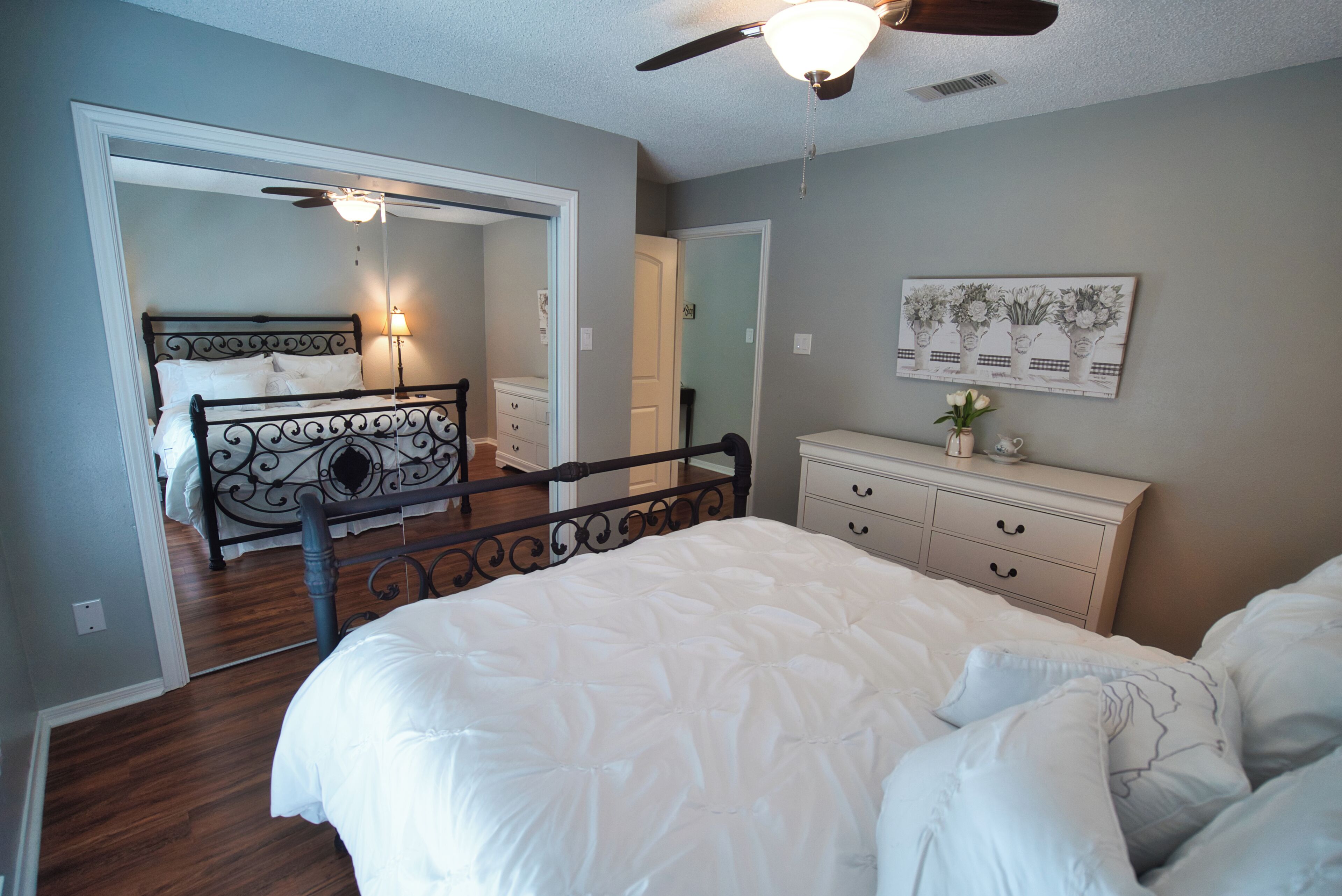A bedroom with a bed, a dresser, a ceiling fan, and a door leading to another room.