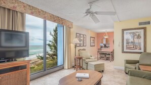 Habitación de hotel con un ventanal grande que ofrece vistas al océano, un televisor, una zona de comedor y un ventilador de techo.