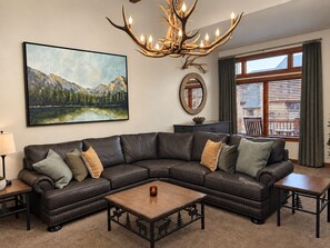 A living room with a large sectional sofa, a coffee table, a chandelier, a painting, and a window with blinds.