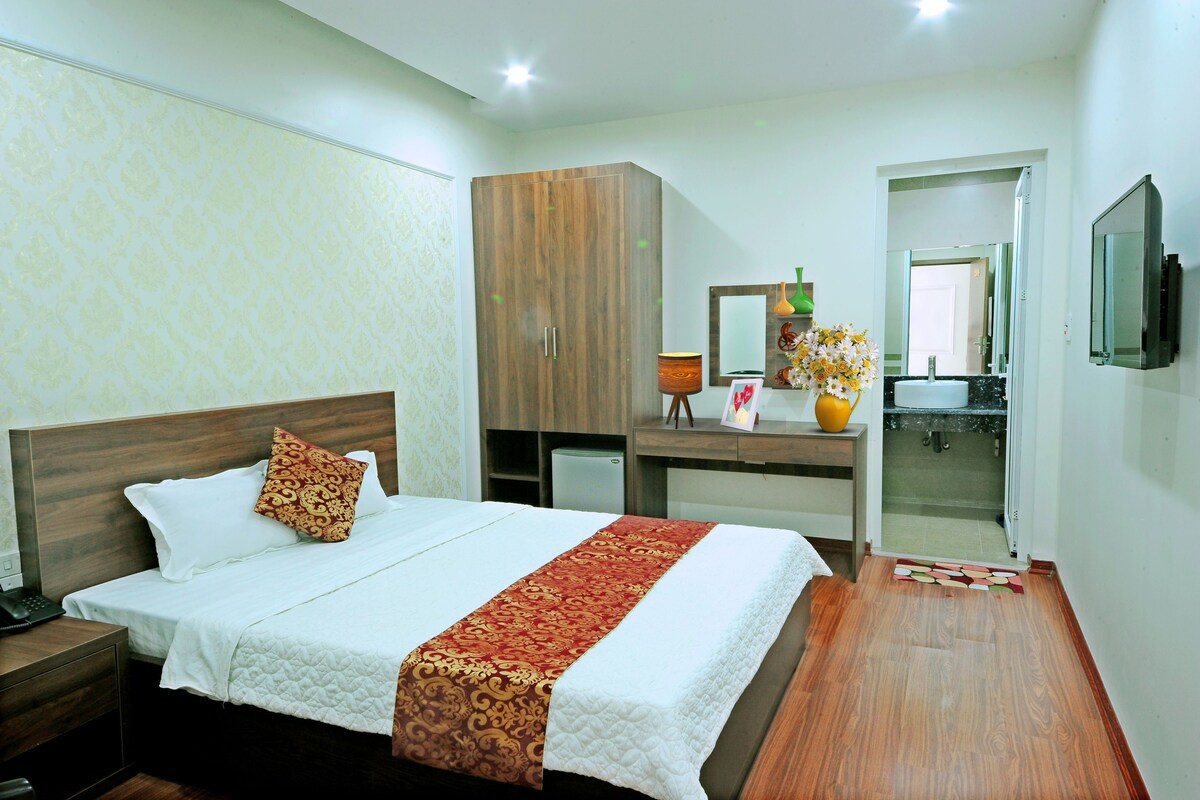 A hotel room with a bed, a wardrobe, a desk with a lamp, a vase with flowers, and a television.