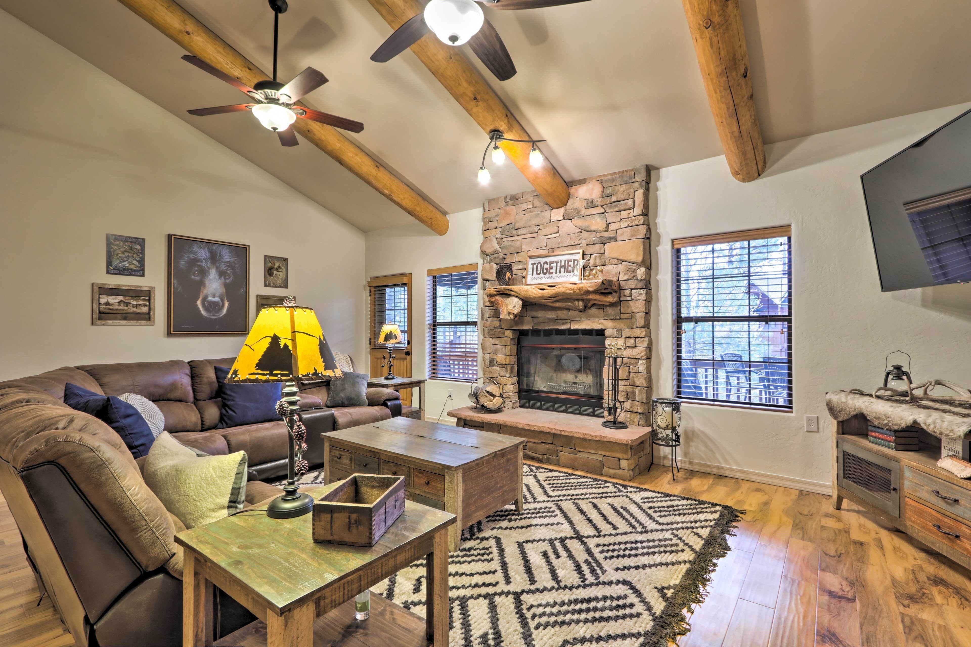 A cozy living room with a stone fireplace, wooden beams, a ceiling fan, and a large window.