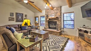 A cozy living room with a stone fireplace, wooden beams, a ceiling fan, and a large window. - Furnished Balcony w/ Fire Pit: Pinetop Mtn Escape! (Pinetop)