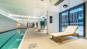 Área interna de piscina moderna, com paredes de vidro, espreguiçadeiras de madeira e piso revestido.