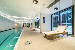 A modern indoor pool area with clear glass walls, wooden loungers, and a tiled floor.