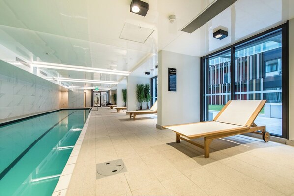 A modern indoor pool area with clear glass walls, wooden loungers, and a tiled floor. - Designed Penthouse 2Bed & 2Bath with SkyTower View on Queen St (Auckland)
