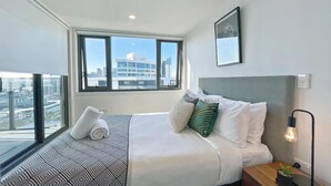 Quarto de hotel moderno com uma cama grande, vista da cidade e paisagem urbana visível pela janela.