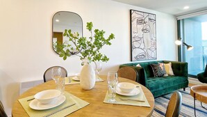 Espaço de refeições com mesa redonda posta para dois, um vaso com folhagem verde e um sofá verde ao fundo.