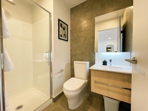 A modern bathroom with a glass shower enclosure, a white toilet, and a wooden vanity with a mirror. - Designed Penthouse 2Bed & 2Bath with SkyTower View on Queen St (Auckland)